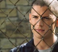 a young man behind a chain link fence looking at the camera with an intense look on his face