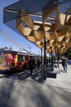 people are walking on the sidewalk next to a bus stop with an artistic wooden structure