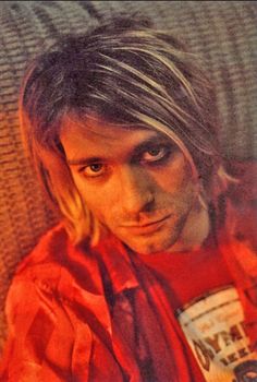 a man with long hair wearing a red shirt and looking at the camera while sitting on a couch