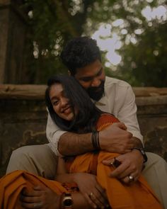 a man and woman hugging each other while sitting on a bench in front of trees