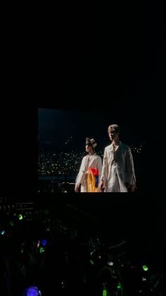 two people standing on a stage in front of a large screen with green and yellow lights