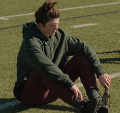 a young man sitting on the ground with his foot up in front of him while wearing a green sweatshirt and maroon sweatpants