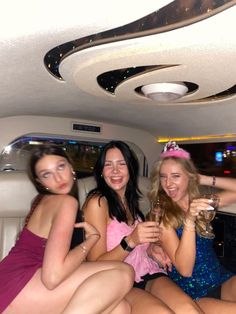 three women in dresses sitting on the back of a car holding champagne flutes and smiling