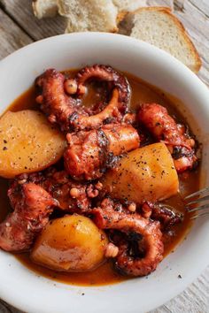 a white bowl filled with meat and potatoes on top of a wooden table next to bread