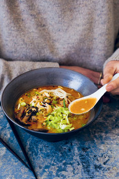 a person holding a spoon over a bowl of soup with noodles and vegetables in it