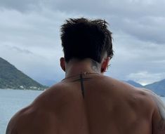 a man with a cross tattoo on his back looking out at the water and mountains