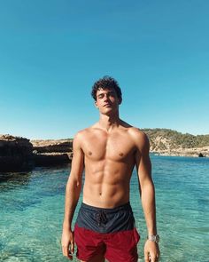 a shirtless man standing in shallow water near the ocean with his hands on his hips