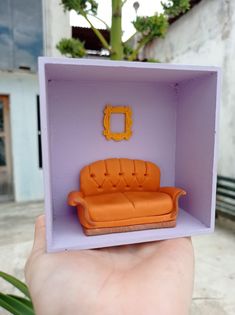 someone is holding up a miniature couch in a tiny purple box with an orange sofa inside