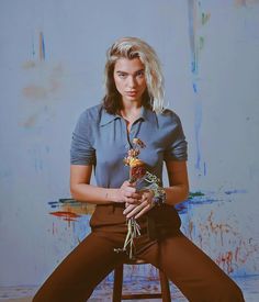 a woman sitting on top of a stool holding a flower in her right hand and looking at the camera