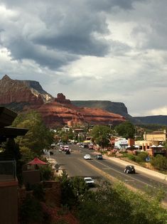 cars are driving down the road in front of some red rocks and green trees under a cloudy sky