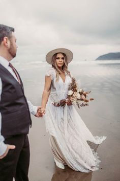 a woman in a white dress and hat walking on the beach holding hands with a man