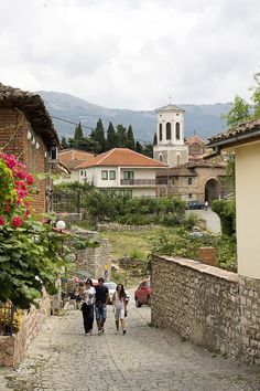 three people walking down a cobblestone street in an old european village with mountains in the background