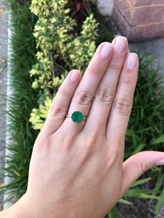 a woman's hand with a green ring on her finger and some plants in the background