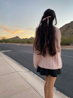 a woman standing on the sidewalk with her back to the camera, wearing a pink sweater and black skirt