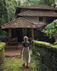 a woman walking down a dirt path past a building with trees and bushes on both sides