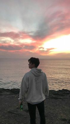 a young man standing on top of a rocky beach next to the ocean at sunset