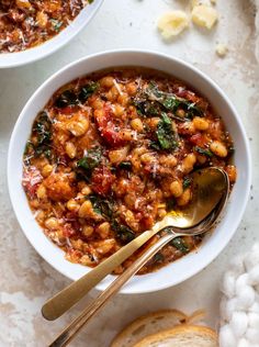 two bowls filled with pasta and spinach on top of a white table next to bread