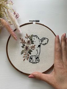 a hand is holding a cross - stitch embroidery hoop with a cow and flowers on it