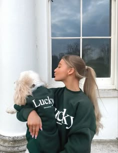 a woman in a green sweatshirt holding a white dog and looking out the window with cloudy sky behind her