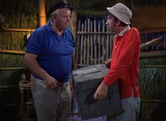 two men standing next to each other in front of a bamboo hut with an open trunk