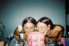 two women leaning over a table with a pink cake in front of them and wine glasses on the table