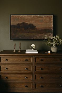 a painting hangs on the wall above a dresser with flowers and candles in front of it