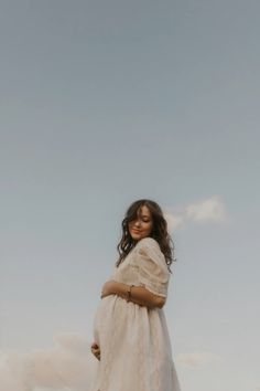 a woman in a white dress standing on top of a grass covered field with clouds