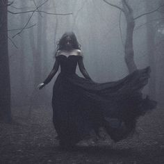 a woman in a long black dress is walking through the woods with her hair blowing in the wind