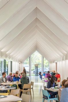 people are sitting at tables in a large room with white walls and ceilings, while one person is standing up to the ceiling