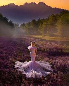 a woman in a white dress standing in a field with lavender flowers and mountains behind her