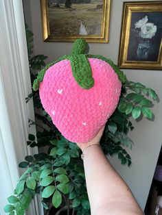 a hand holding up a crocheted strawberry pillow in front of a potted plant