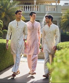 three men in white and pink outfits walking down a sidewalk near a fountain with water sprinkles on it