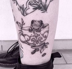 a black and white photo of a frog in a teacup tattoo on the leg