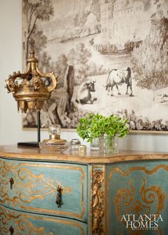 an ornate painted dresser with green plants on top in front of a large wall painting