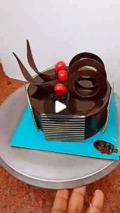 a hand holding a cake with chocolate icing and cherries on top, in front of a white wall