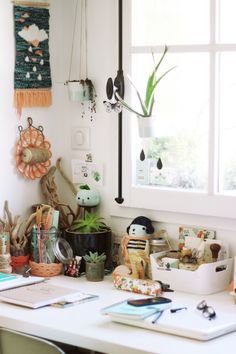 a white desk topped with lots of clutter next to a window filled with potted plants