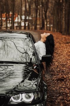 a woman leaning on the hood of a black car in front of trees and leaves