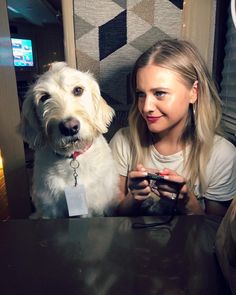 a woman sitting at a table next to a white dog with a tag on it's collar