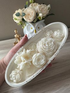 a hand holding a white tray with flowers in it on a table next to a vase