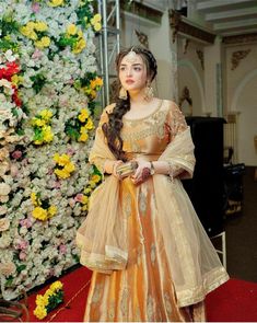 a woman standing in front of a flower wall wearing a yellow dress and gold jewelry