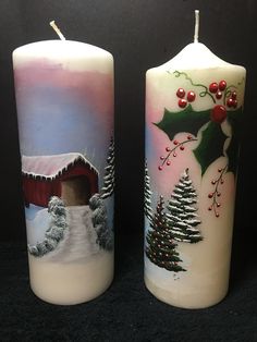 two lit candles decorated with christmas scenes and holly berries on the top one has a red barn in the background