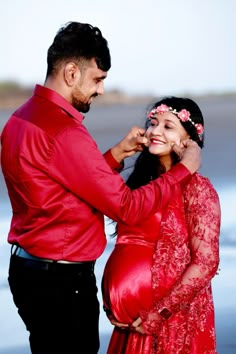 a pregnant woman in a red dress is fixing her husband's tie at the beach