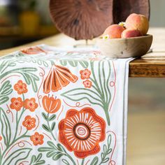 an orange and green floral table runner next to a bowl of peaches on a wooden table