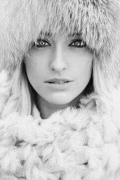 a woman with blue eyes wearing a fur hat and scarf over her head is looking at the camera