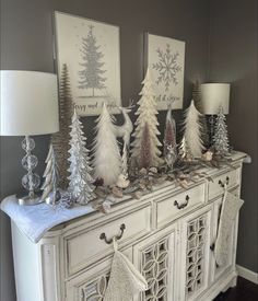 a white dresser topped with christmas trees and stockings on it's sideboard next to two lamps