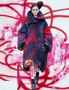 a woman is walking down the runway wearing a colorful coat and black tights, with graffiti on the wall behind her