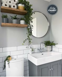 a white toilet sitting next to a bathroom sink under a round mirror and wooden shelves