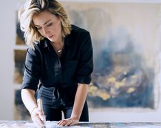 a woman in black shirt and pants painting on large piece of paper with paintbrush