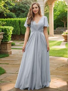 a woman in a long grey dress standing on a stone walkway with trees and bushes behind her