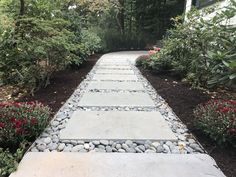 a stone path in the middle of a garden with flowers and trees around it, leading to a house